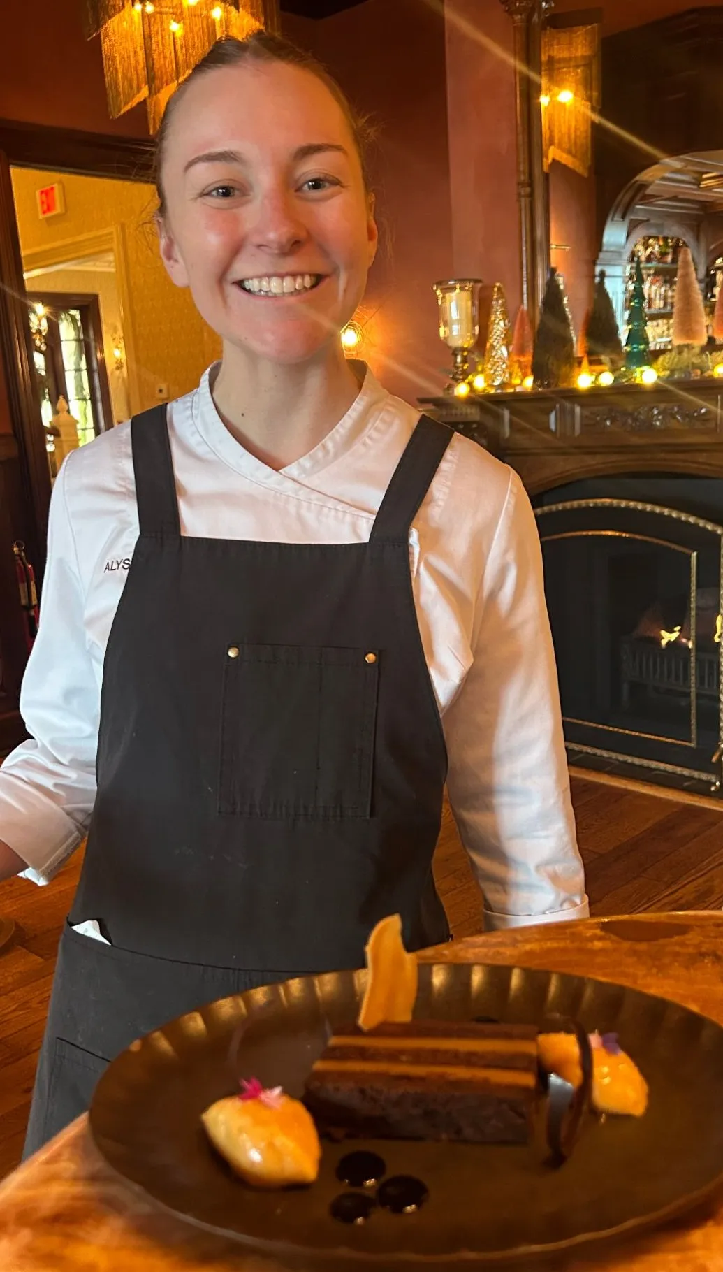 Alyssa May in chef whites and a black apron, smiling behind a plated layered chocolate dessert