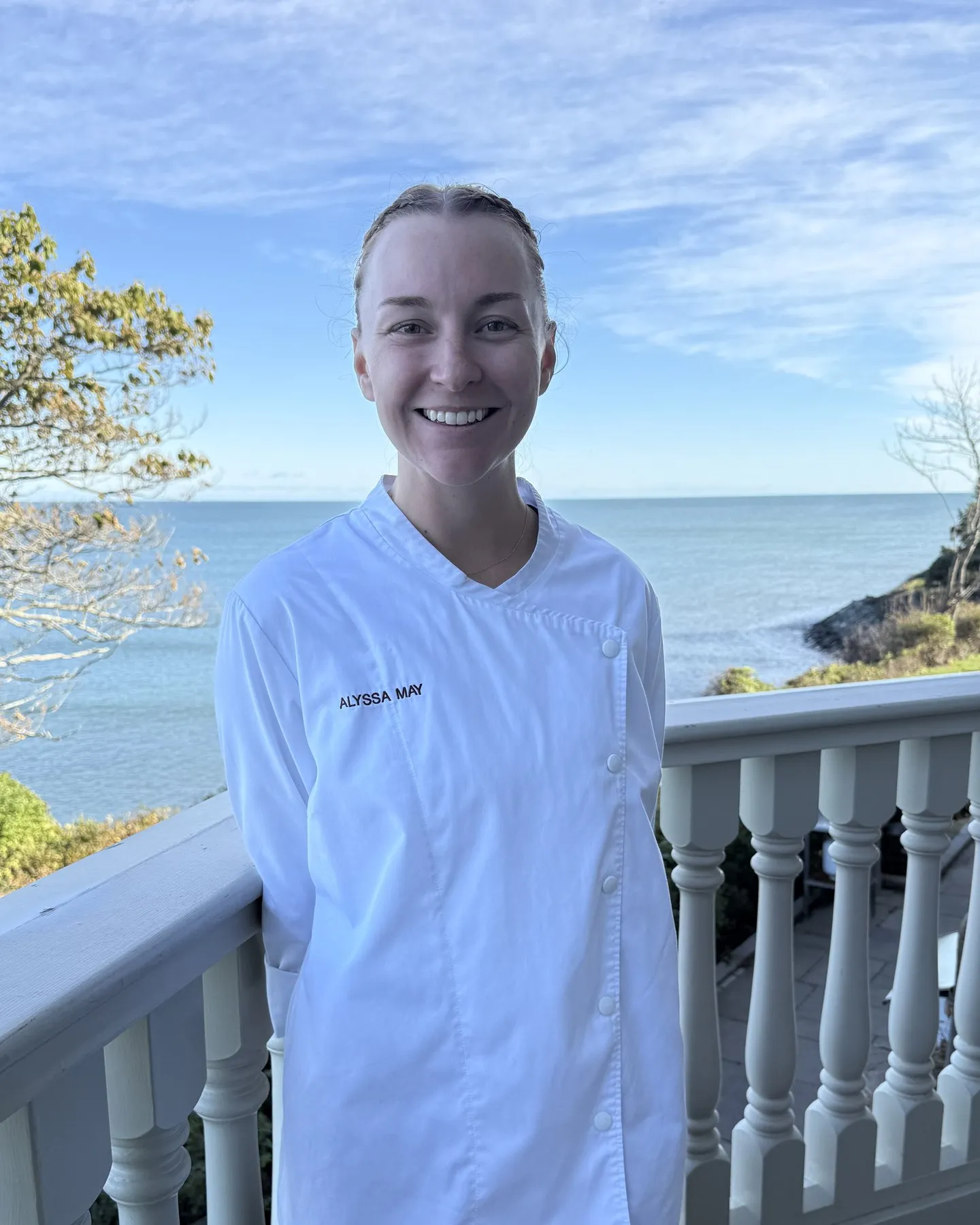 Alyssa May in chef whites, smiling on a porch with the ocean behind her