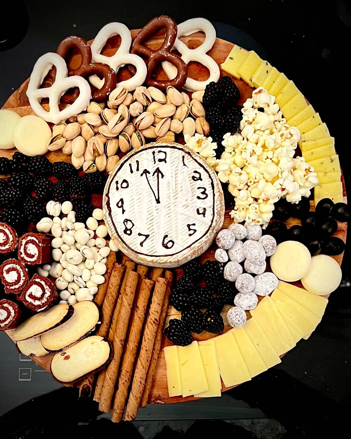 New Year's Eve board with a clock drawn on a wheel of brie, surrounded by chocolate pretzels, pistachios, blackberries, popcorn, and cheese