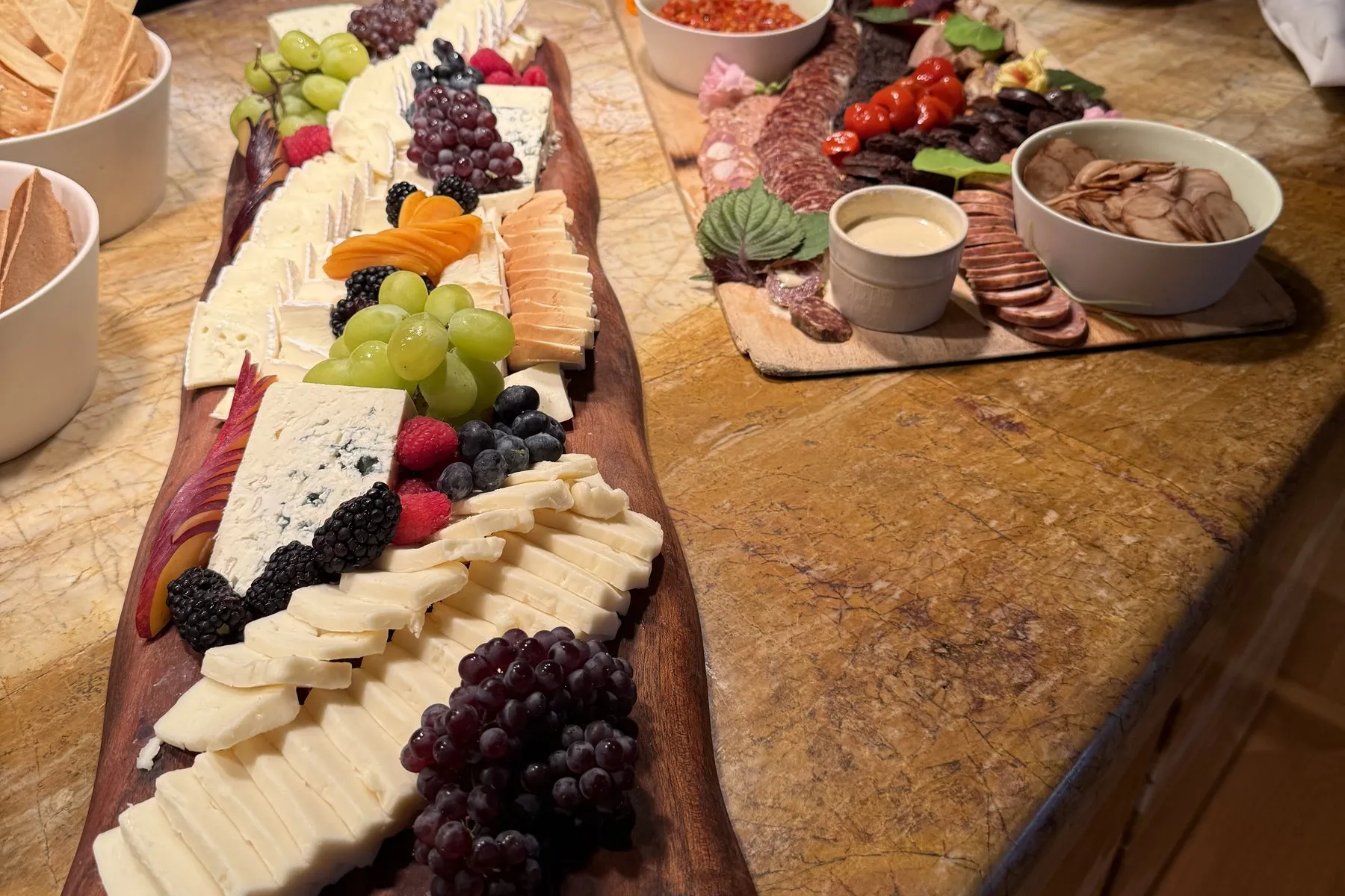 Long wooden grazing board of sliced cheeses, blue cheese, grapes, and blackberries on a kitchen island