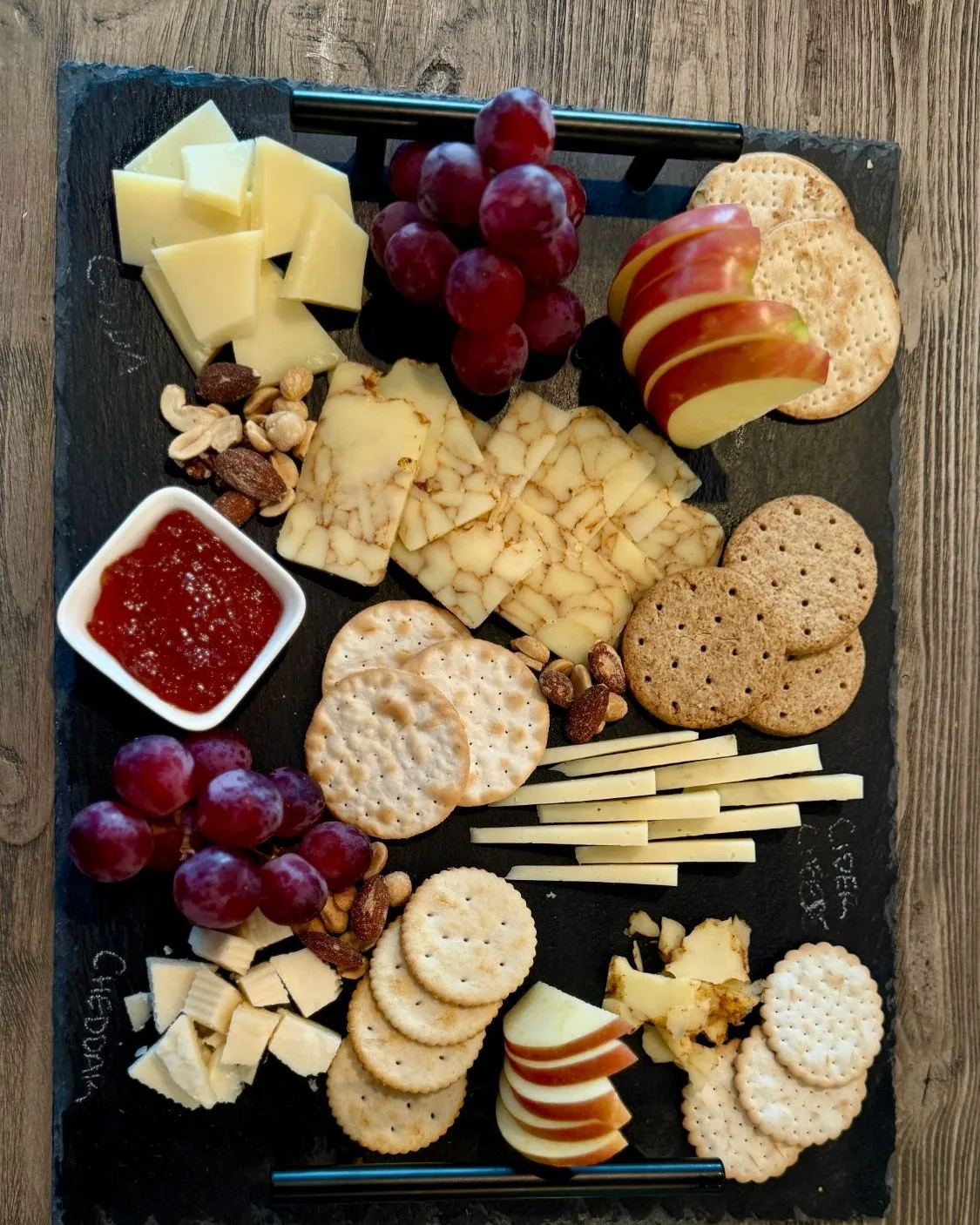 Slate cheese board with chalk-labeled gouda and cheddar, grapes, apple slices, crackers, almonds, and a dish of jam