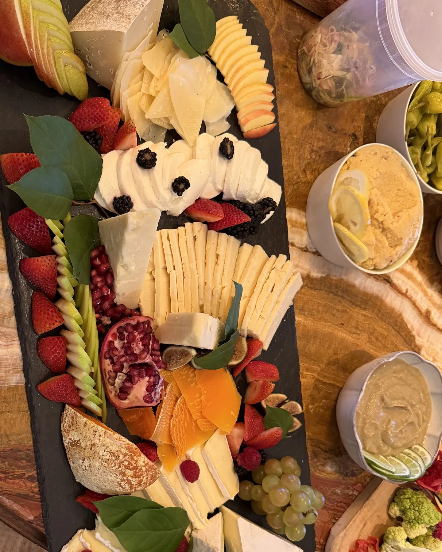 Long slate board of soft and hard cheeses with strawberries, pomegranate, orange, and dips on the side